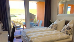 A double bed with white bedding and floral-patterned pillows, a small bedside table, and a window with a view of outdoor seating. - Residenz Windjammer (Cuxhaven)