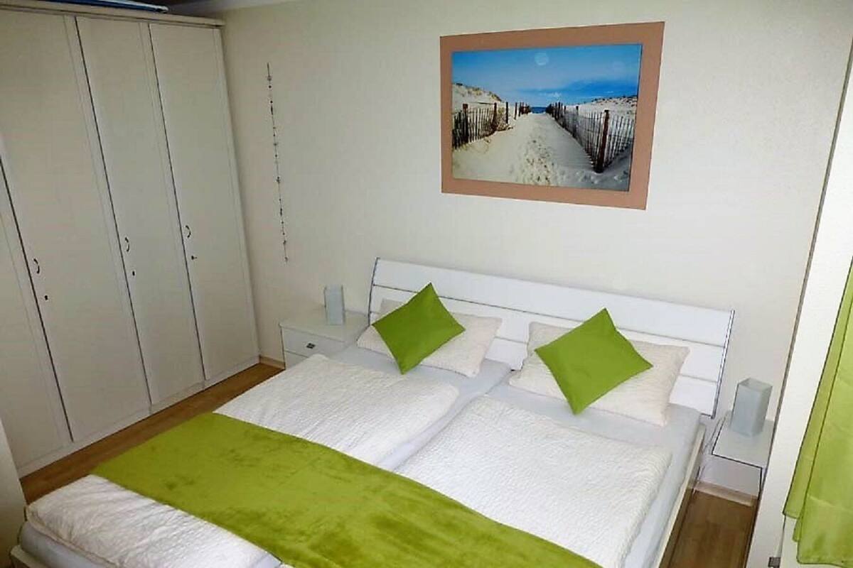 A bedroom with a bed, green pillows, a white headboard, a nightstand, a wall-mounted painting, and a wardrobe.