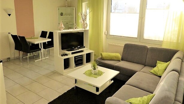 A living room with a grey sofa, a white coffee table, a TV stand, and a dining area with a table and chairs. - Haus Seemöwe (Cuxhaven)
