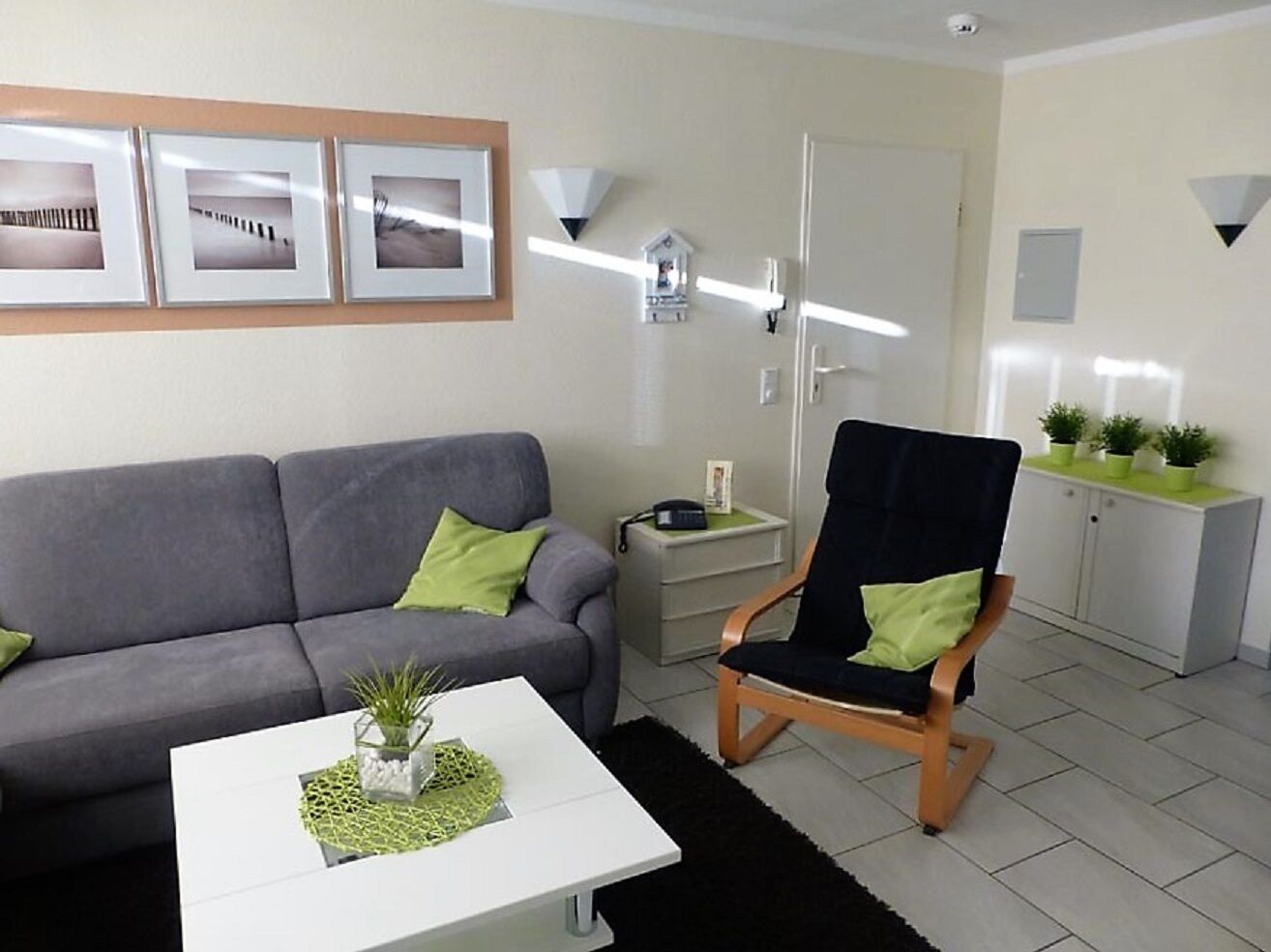 Un salon avec un canapé gris, une chaise en bois, une table basse blanche ornée d’une plante, et un meuble sur lequel sont disposées des plantes en pot.