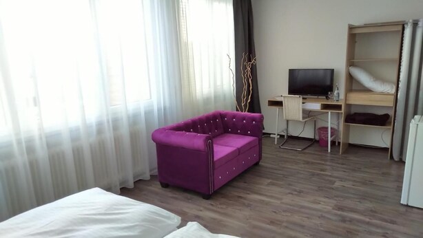 A hotel room with a wooden floor, a purple sofa, a bed with white linens, a desk with a computer and a TV, and a wardrobe. - Atelier Guesthouse (Filderstadt)