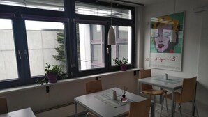A modern dining area with tables and chairs, large windows with potted plants, and a poster on the wall. - Atelier Guesthouse (Filderstadt)