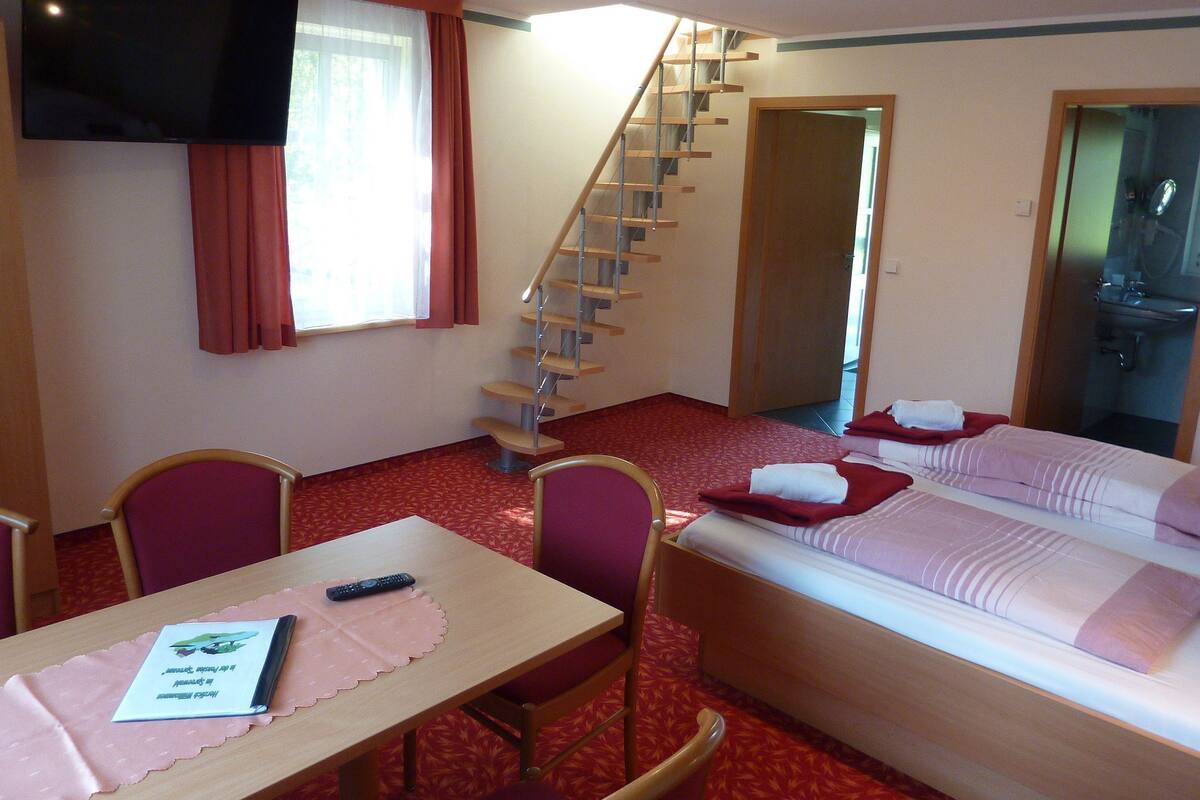 Ein Hotelzimmer mit Bett, Schreibtisch mit Fernbedienung, Stuhl, Treppe und einem durchgegangene Tür sichtbaren Badezimmer.
