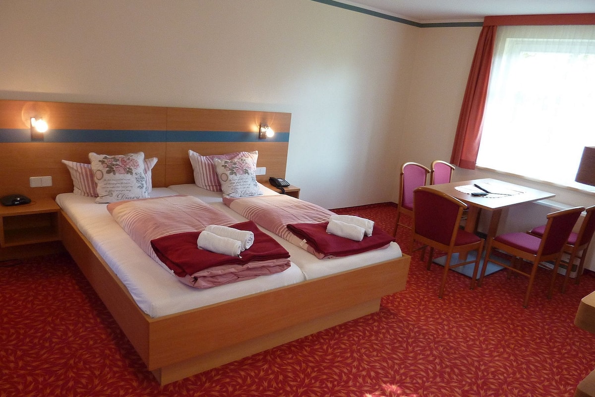 Ein Hotelzimmer mit einem großen Bett, zwei Nachttischen, einem Schreibtisch mit Stuhl und einem fensterbepflanzten Fenster.