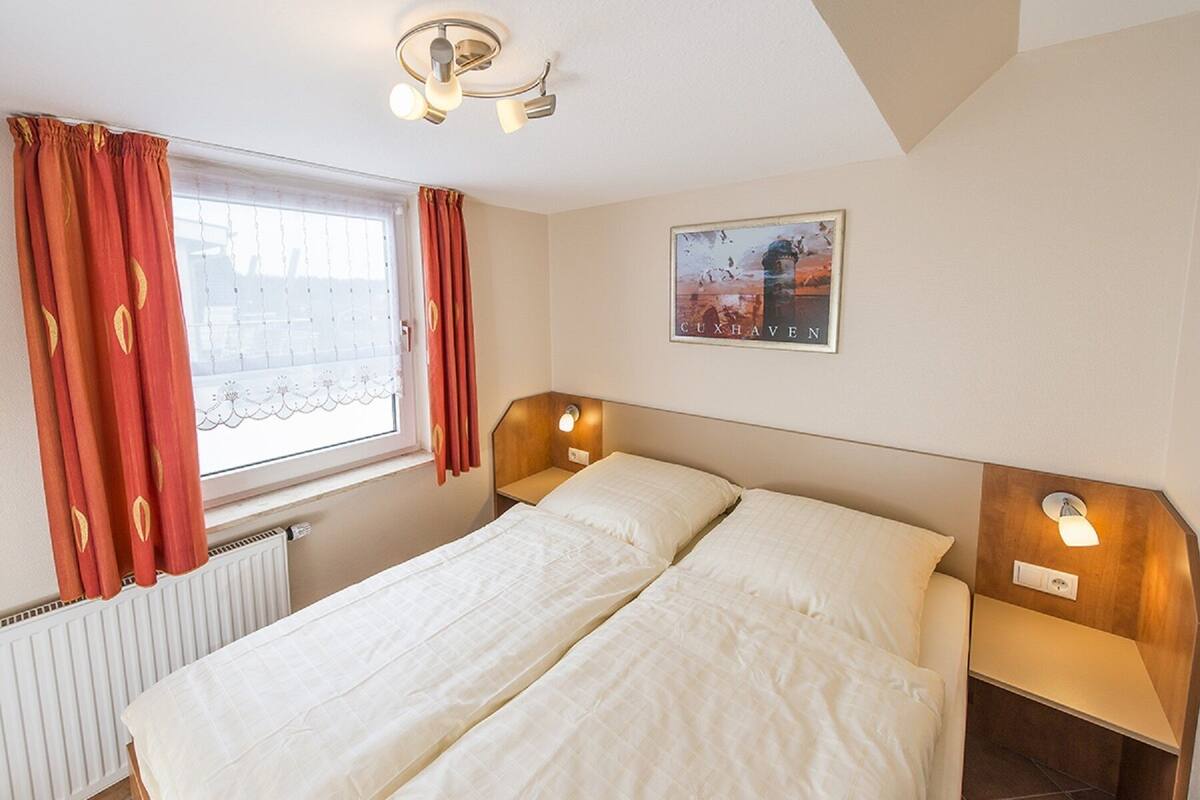 A hotel room with a bed, two bedside lamps, a window with curtains, a radiator, and a framed picture on the wall.