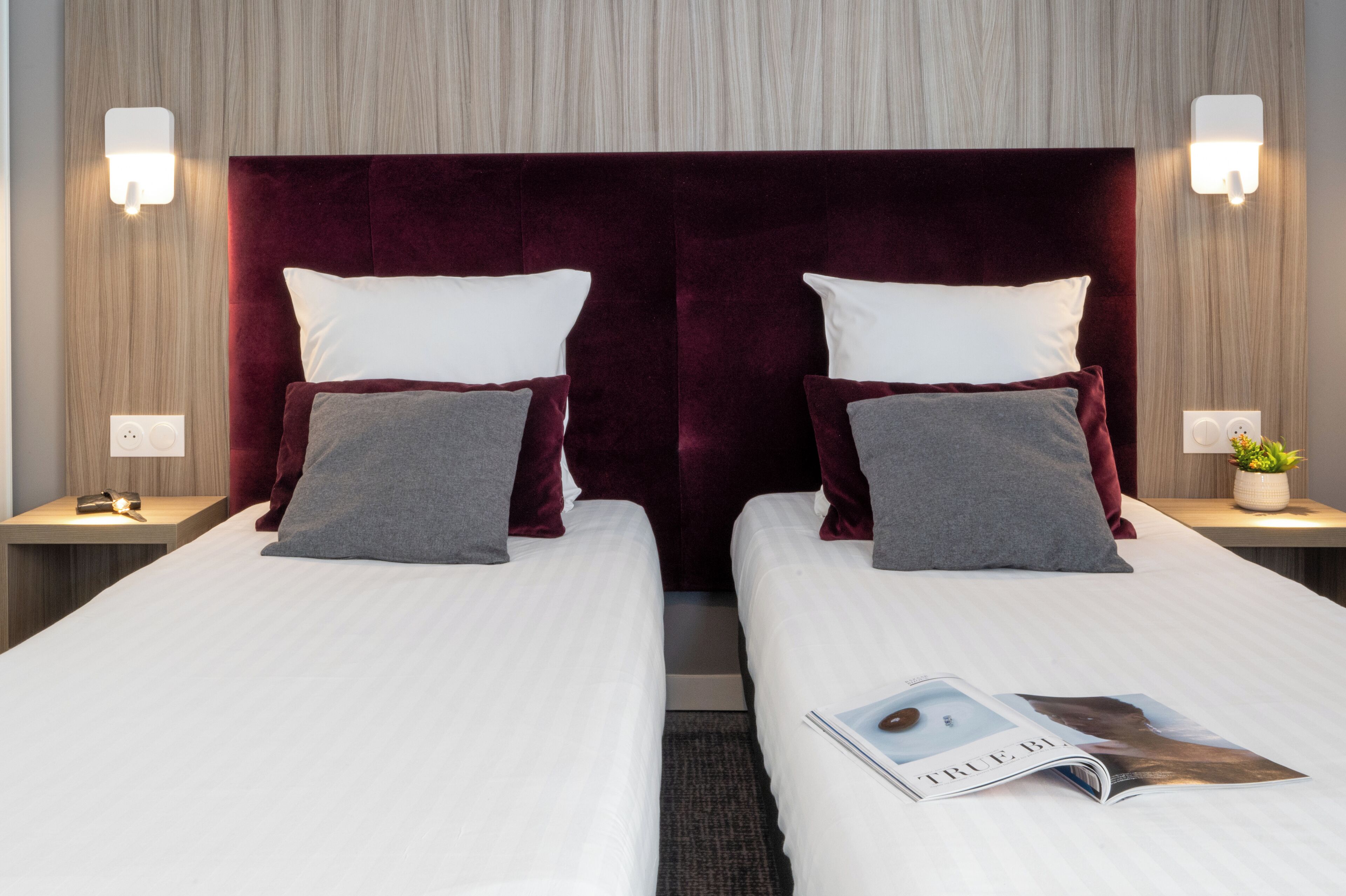 A hotel room with two beds, each with white bedding and maroon headboards. There are wall-mounted light fixtures on either side of the beds, and a small table with a plant on the right side.