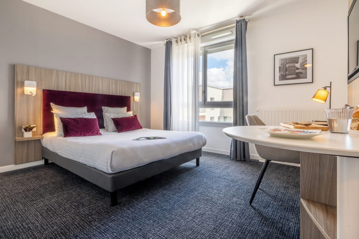 Ein modernes Hotelzimmer mit einem großen Bett, einem Nachttisch, einem Sessel und einem Fenster mit Vorhängen.