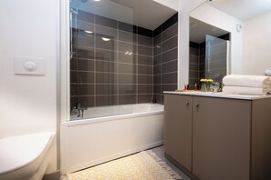 A modern bathroom with a bathtub, a toilet, a vanity with a mirror, and a patterned rug. - Nemea Appart Hotel Grand Cœur Nancy Centre (Nancy)