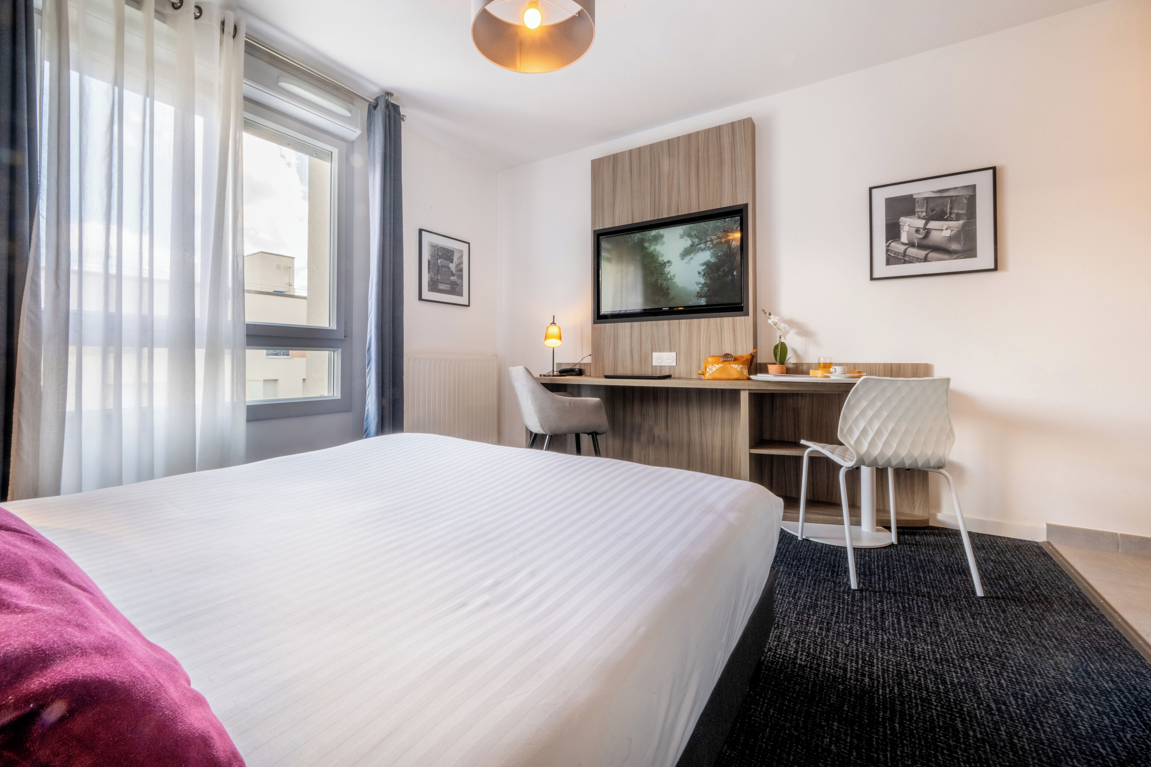 A hotel room with a large bed, a desk with a computer, a chair, a TV, and a window with curtains.