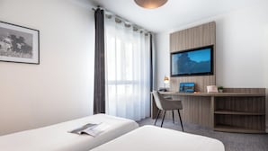 Hotellihuone, jossa on kaksi sänkyä, työpöytä tietokoneella, televisio ja seinällä oleva kehystetty kuva.