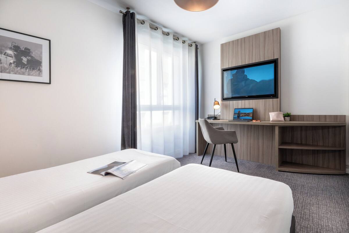 Ein Hotelzimmer mit zwei Betten, einem Schreibtisch mit Computer, einem Fernseher und einem gerahmten Bild an der Wand.
