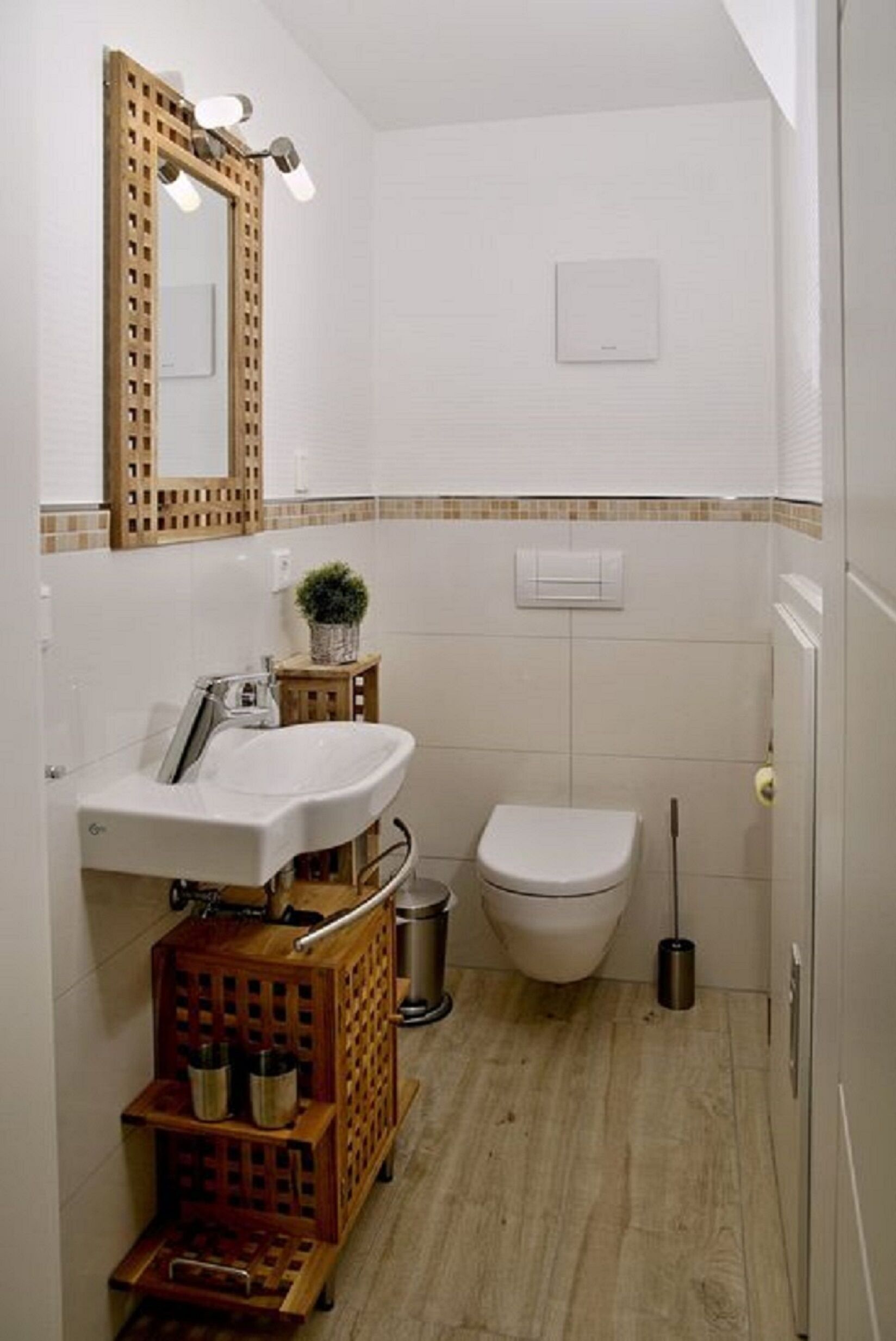 A compact bathroom with a white sink, a toilet, a wooden cabinet, a mirror, and a small plant.