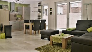 Ein modernes Wohnzimmer mit Sofa, Hocker und Couchtisch. Es gibt einen Essbereich mit Stühlen und Tisch. Der Raum hat große Fenster und einen Ausblick nach draußen.