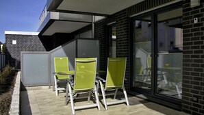 Eine Terrasse mit drei grünen Stühlen und einem Tisch, ein modernes Gebäude mit großen Fenstern und eine Ziegelwand.