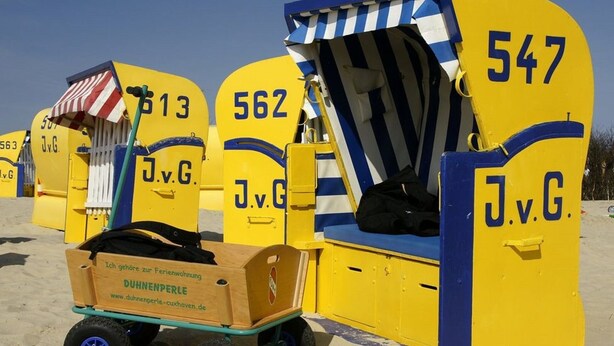 Strandhütten mit den Nummern 547, 562 und 563 in Blau und Gelb, vor ihnen ein Holzwagen mit der Aufschrift 'DUHNNENPERLE'.