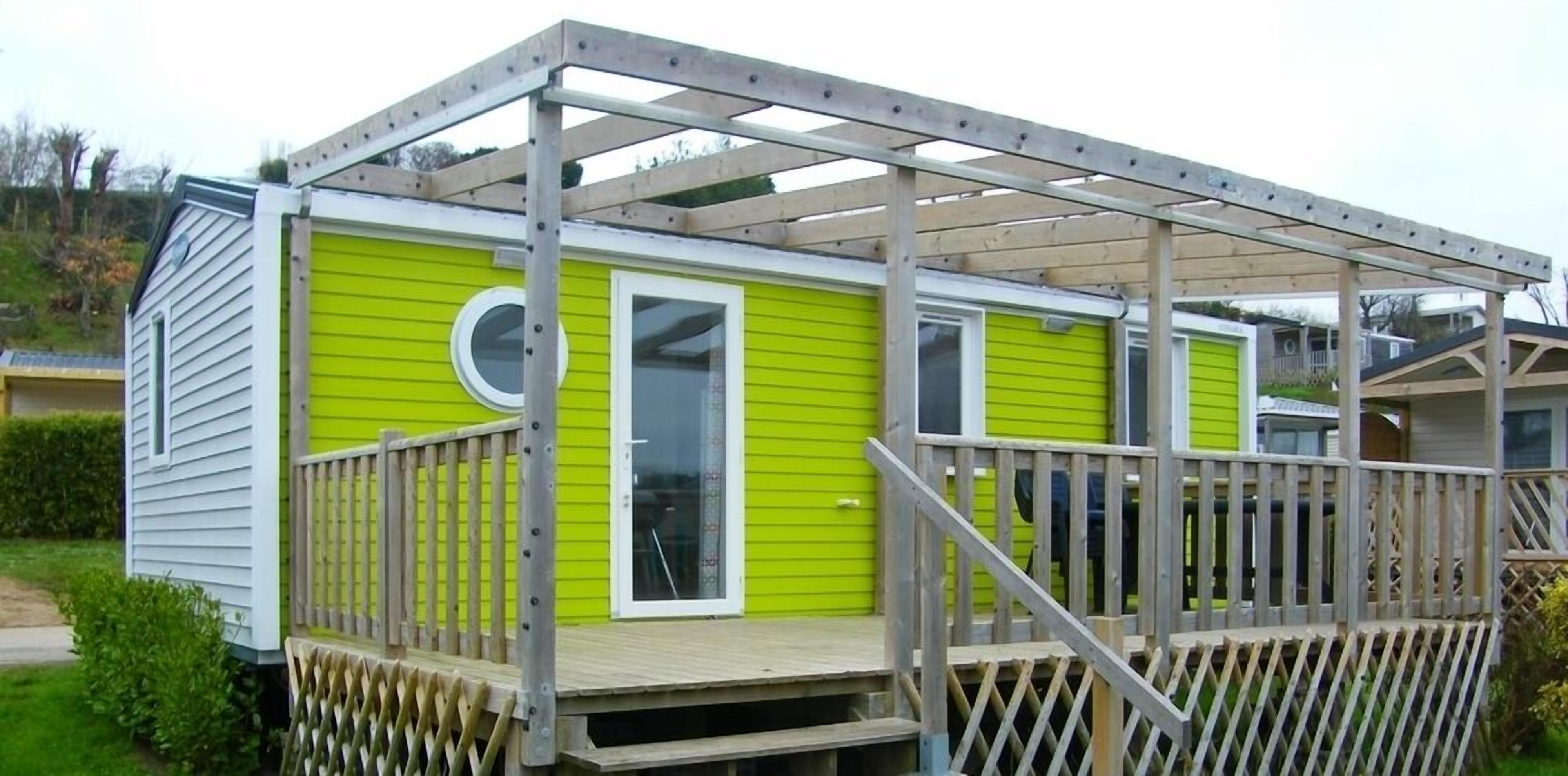 Uma casa pequena, térrea, com fachada verde, deck de madeira e varanda coberta.
