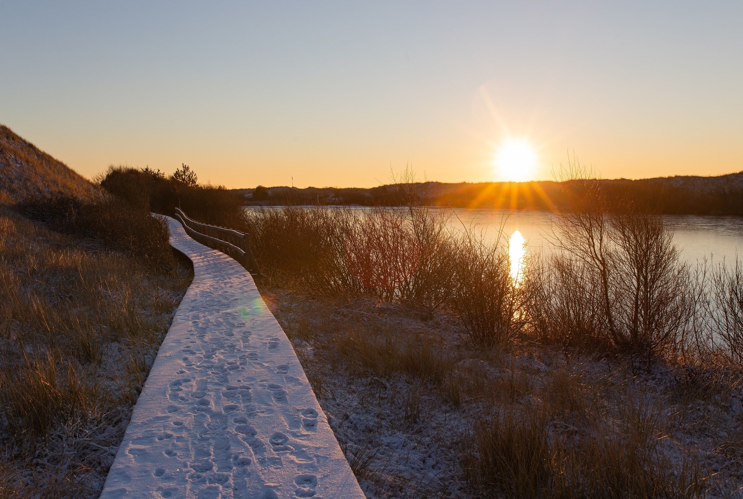 A snow-covered path leading to a body of water with the sun setting in the background.