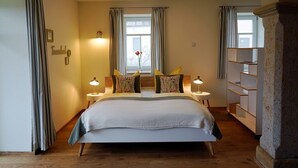 A bedroom with a large bed, two bedside tables with lamps, a window with curtains, and a bookshelf. - Ferienhof Gräfe - Apartment "Spring meadow" with terrace, pool & sauna (Meißen)