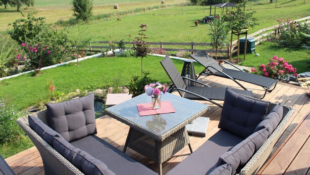 A deck with wicker furniture, a glass table, and a view of a grassy field with trees and a wooden fence. - Ferienhof Gräfe - Apartment "Spring meadow" with terrace, pool & sauna (Meißen)