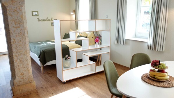 Une chambre à coucher moderne avec un lit, un meuble de rangement blanc, une table à manger avec des fruits, et une vue sur l’extérieur.