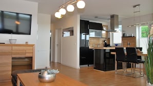 A modern kitchen with a dining area, a TV mounted on the wall, and a large window providing natural light. - Beachhouse (Cuxhaven)
