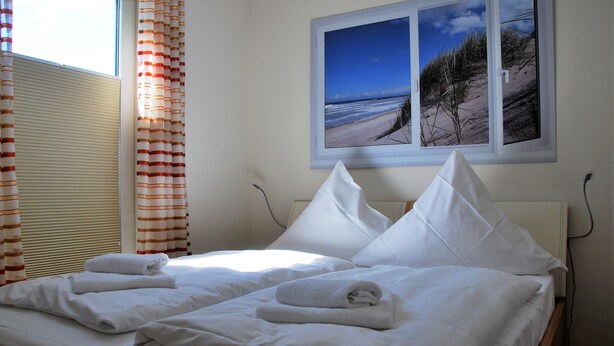 A neatly made bed with white linens and towels, a window with a beach scene, and striped curtains. - Beachhouse (Cuxhaven)