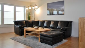 A modern living room with a sectional sofa, a coffee table, and a large window with blinds. - Beachhouse (Cuxhaven)