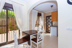 A dining area with a table and chairs, a kitchen in the background, and a view through a glass door.