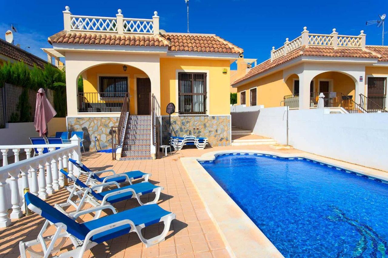 A pool area with lounge chairs, a tiled deck, and a building with a tiled roof and white balcony railings.