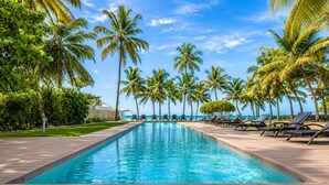 A rectangular swimming pool with lounge chairs and palm trees, surrounded by a wooden deck. - Beachfront | Ocean View Balcony | Pool & Jacuzzi (Playa Juan Dolio)