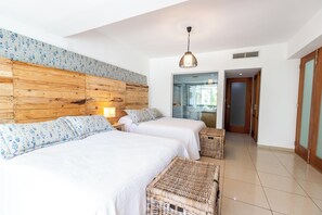 Camera d'albergo con due letti, una testiera in legno, una panca in vimini e un'ampia finestra.
