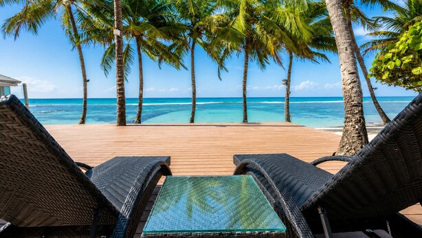 A wooden deck with wicker lounge chairs and a glass-top table overlooking a beach with palm trees and clear blue water. - Beachfront | Ocean View Balcony | Pool & Jacuzzi (Playa Juan Dolio)
