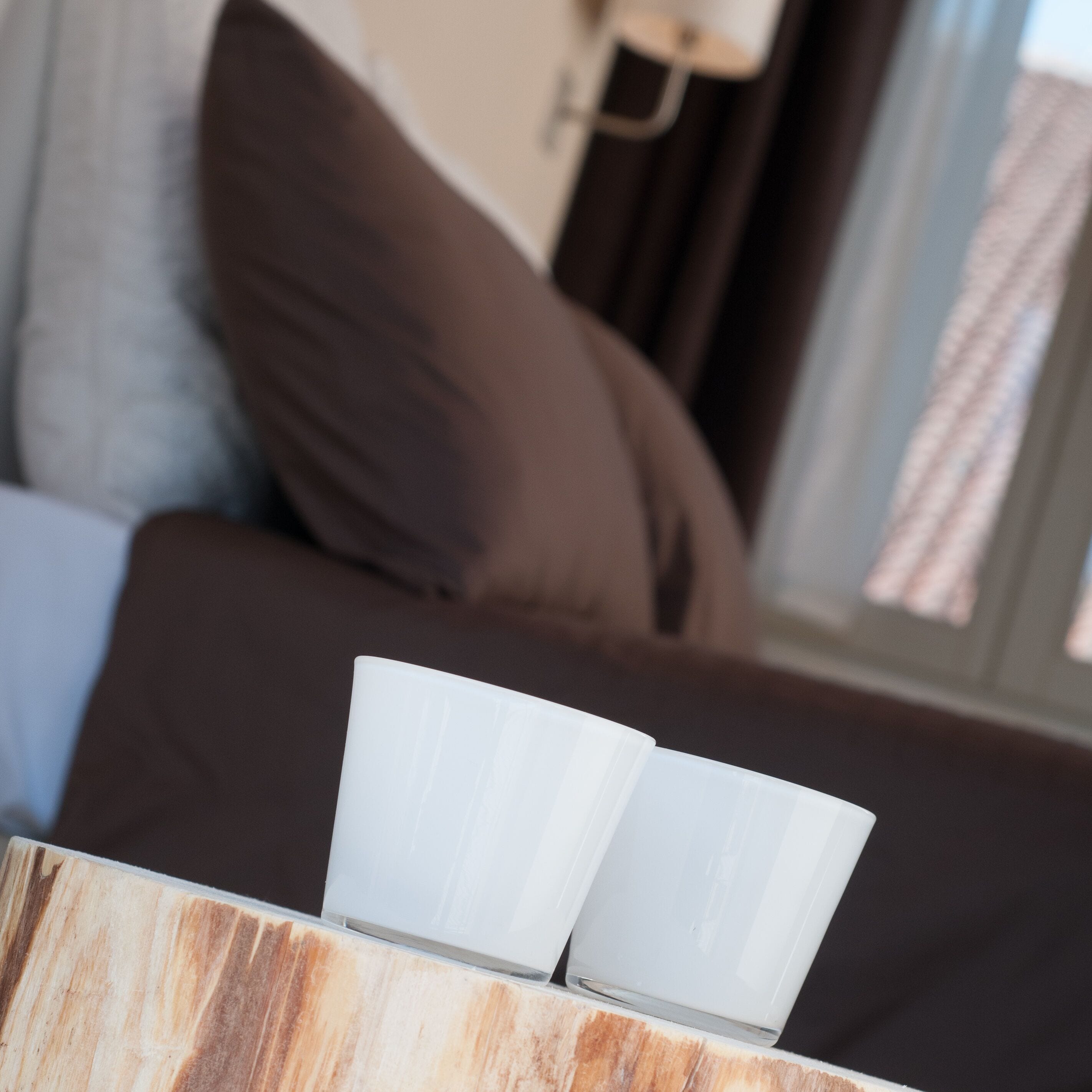 Una mesa de madera con dos tazas blancas encima, frente a un sofá marrón con una ventana al fondo.