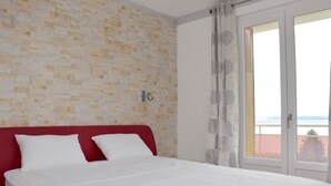 Un dormitorio con cabecera roja, cubrecama blanco y vista al mar a través de un ventanal grande.