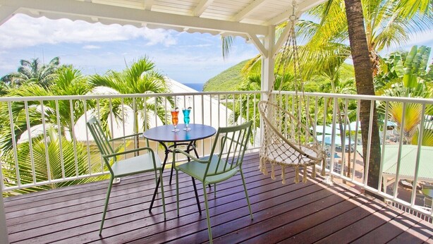 A covered outdoor area with a wooden deck, two green chairs, a small round table, and a hanging hammock.