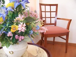 A wooden chair with a cushioned seat and backrest, positioned next to a white vase with a textured design containing a floral arrangement. - Mayna (Benidorm)