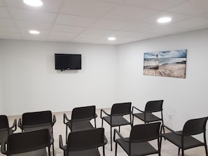 A conference room with a flat-screen TV mounted on the wall, a beach-themed painting, and rows of black chairs arranged for seating. - Mayna (Benidorm)