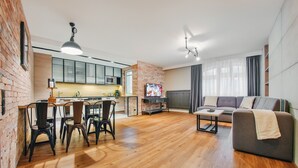 A modern living room with a wooden floor, a grey sofa, a dining area with chairs, a kitchen with a brick wall, and a TV on the wall. - Apartamenty Sun & Snow Centrum - Plaża (Sopot)