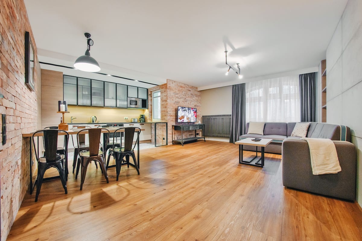 Een moderne woonkamer met een houten vloer, een grijze bank, een eethoek met stoelen, een keuken met een bakstenen muur, en een televisie aan de muur.