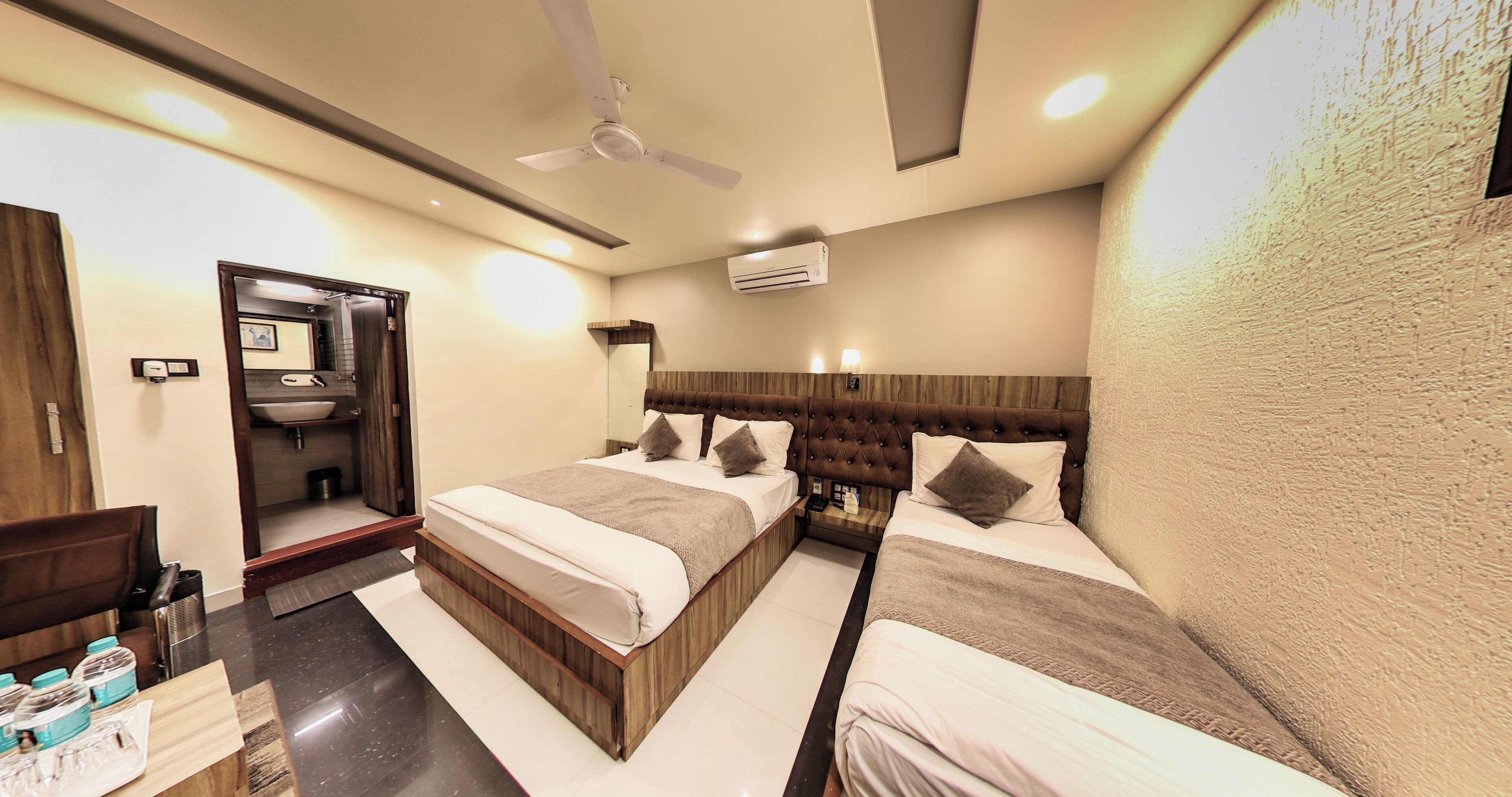Une chambre d’hôtel avec deux lits, un ventilateur de plafond, un appareil de climatisation, une table de chevet avec une lampe et une salle de bain avec un lavabo et un miroir.