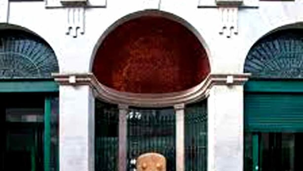 Un edificio con un arco centrale, affiancato da due porte verdi, e una panchina in pietra di fronte.