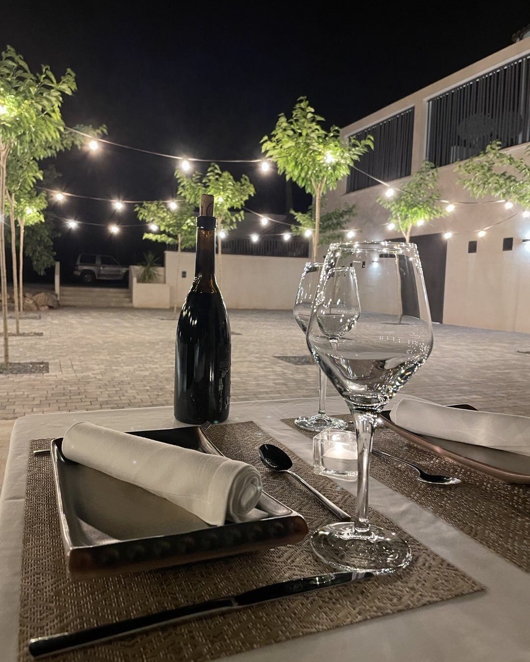 Impostazione esterna per la cena con una bottiglia di vino, due bicchieri e un tavolo ordinato con tovagliette e posate.