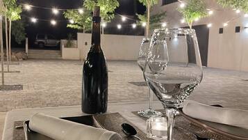 Impostazione esterna per la cena con una bottiglia di vino, due bicchieri e un tavolo ordinato con tovagliette e posate.
