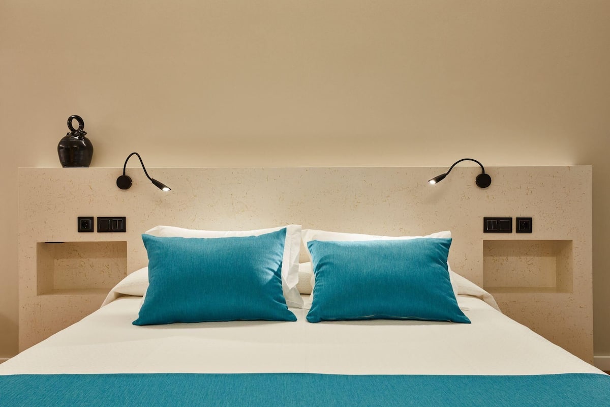 Een netjes opgemaakt bed met twee blauwe kussens en een witte deken. Aan beide zijden van het bed hangen twee wandlampen.