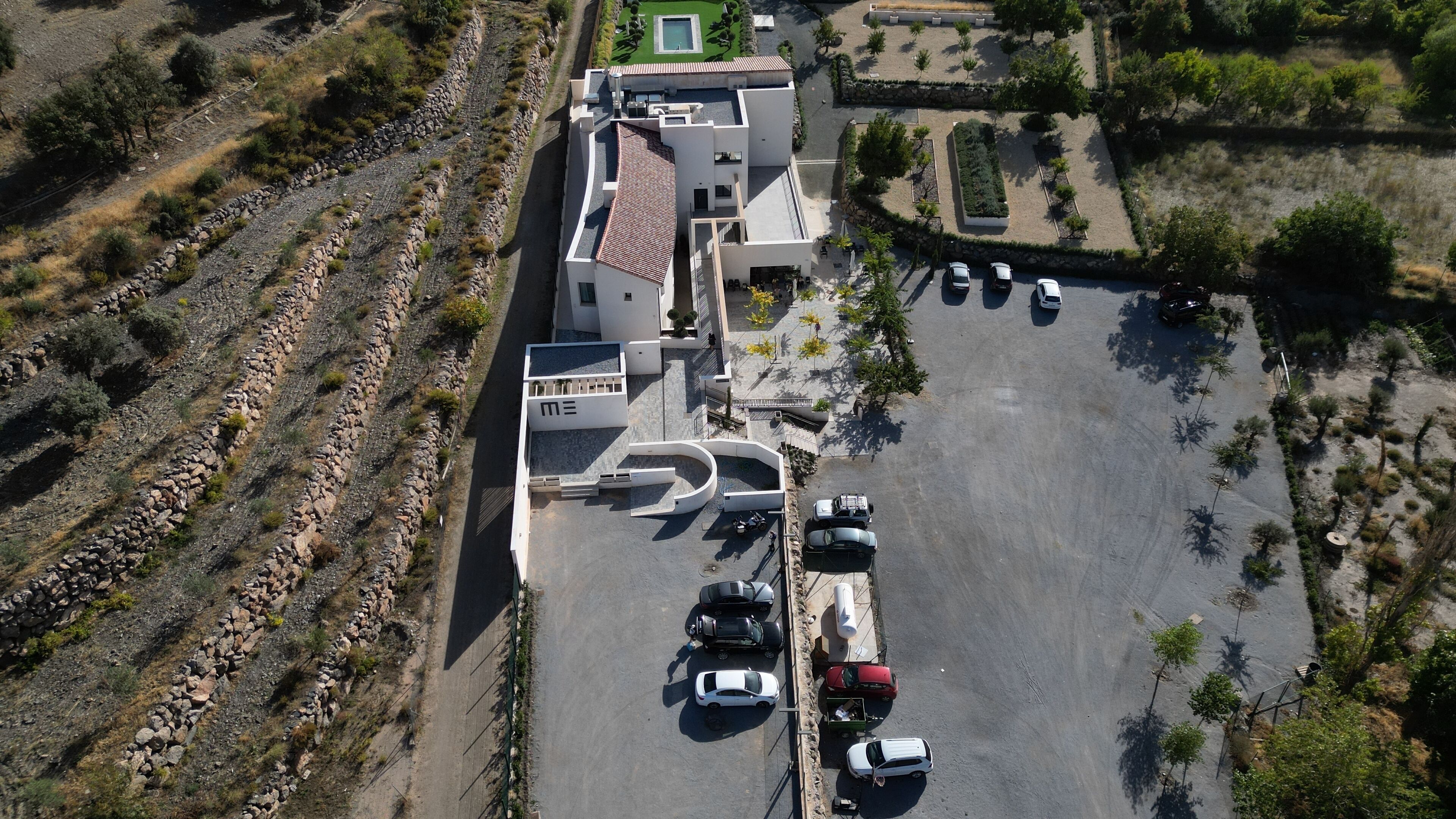 Une vue aérienne d’un parking où se trouvent plusieurs voitures, d’un bâtiment doté d’un toit en pente et d’un espace paysager agrémenté d’arbres et d’arbustes.
