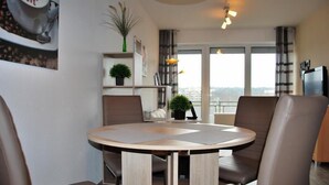 A modern dining area with a round table and four chairs, a bookshelf with decorative items, and a view of the city through large windows. - Haus Seewind 10 (Cuxhaven)