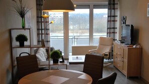 A modern living room with a round table, two chairs, a sofa, a TV, and a balcony with a view of buildings and trees. - Haus Seewind 10 (Cuxhaven)