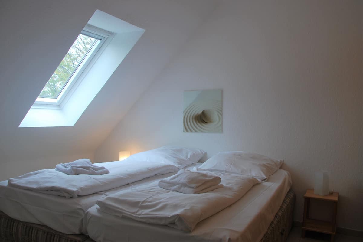 Une chambre avec une lucarne, un lit recouvert de draps blancs, une table de chevet avec une lampe et une œuvre d’art murale.