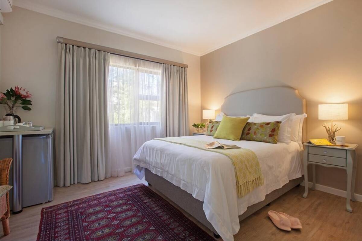 Quarto com cama, mesas de cabeceira, escrivaninha, cadeira e janela com cortinas.