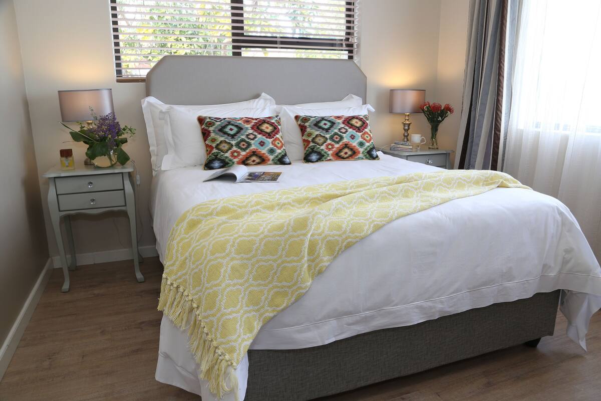 Cama bem arrumada com manta amarela estampada, dois travesseiros decorativos, abajur e mesinha de cabeceira com um vaso de flores.